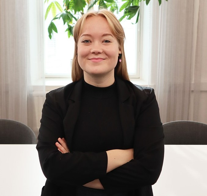 Nainen pukeutuneena mustiin vaatteisiin, hymyilee kädet puuskassa, taustalla viherkasvi ja valoisa ikkuna