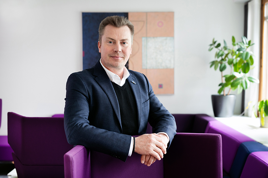 Aki Laiho tummassa pikkutakissa nojaa käsivarsiaan violettien tuolien selkänojiin. Taustalla abstrakti taulu.