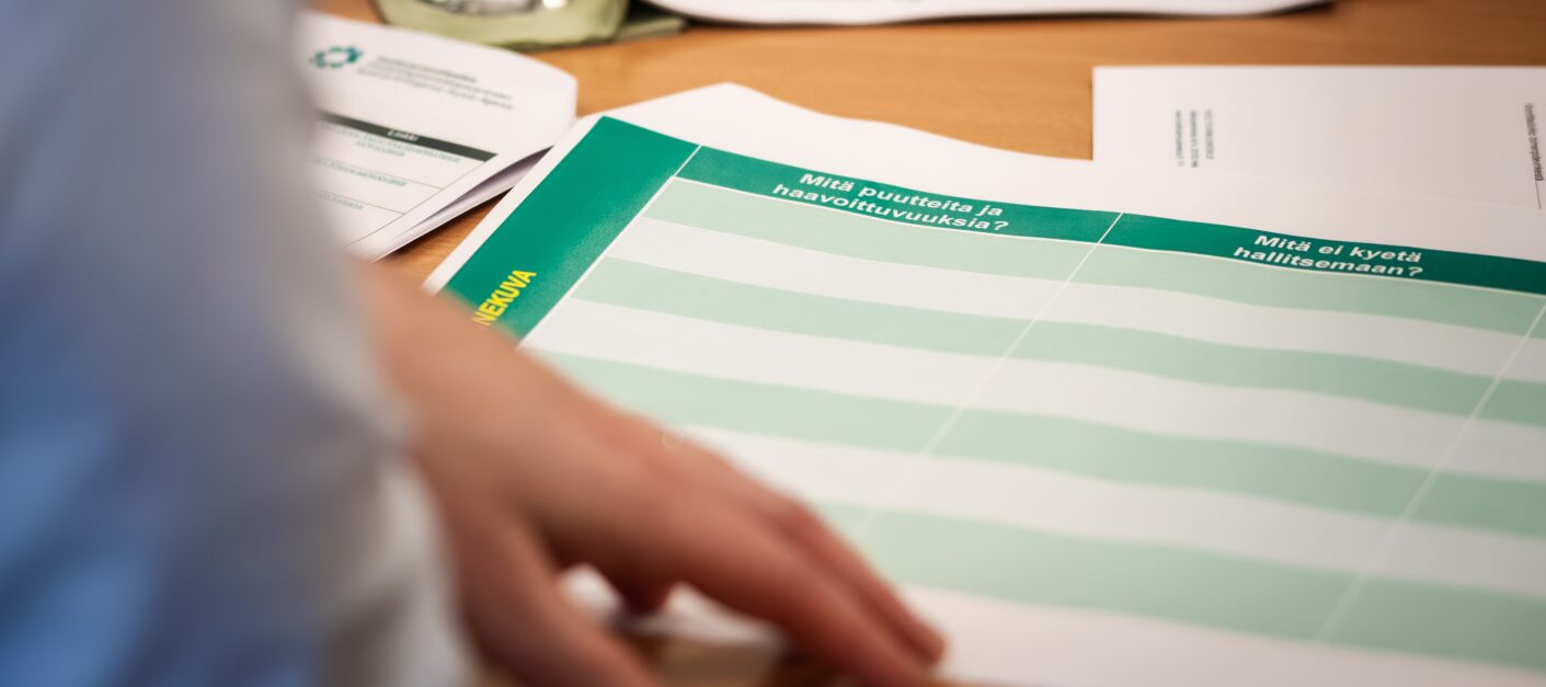 Pöydällä on paperi, jossa lukee omissa sarakkeissaan: "Tilannekuva: mitä puutteita ja mitä haavoittuvuuksia, mitä ei kyetä hallitsemaan." Käsi pitää paperia pöydän päällä.